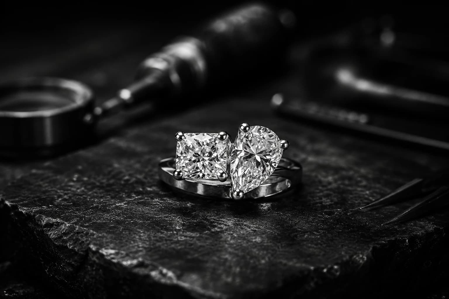 Two bespoke diamond rings — cushion cut and pear shape — photographed on dark stone with jeweller's tools in the background.